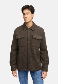 Brunrandig jacka med knäppning fram, två bröstfickor och klassisk krage. Tillverkad av strukturerad tyg, i kombination med blå jeans.