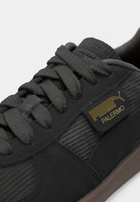 Kratki prikaz crnog Puma Palermo tenisica s teksturiranim vezicama, zlatnom oznakom i smešnim gumiranim đonom.