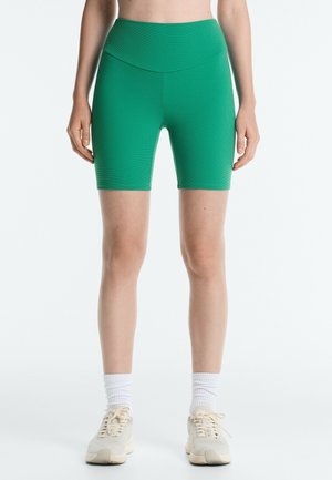 Femme portant un short cycliste vert taille haute, des chaussettes blanches et des baskets beiges, debout devant un fond blanc uni.