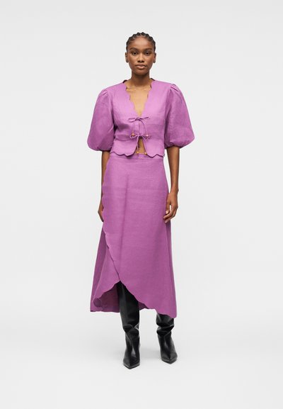 Femme portant un haut violet à manches bouffantes avec des bords festonnés et une jupe portefeuille assortie, associée à des bottes noires montant jusqu'aux genoux.