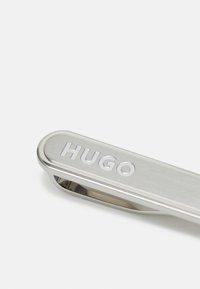 Clip de cravate en argent avec une finition brossée, arborant le mot gravé "HUGO". Design épuré avec des bords arrondis et un fermoir robuste.
