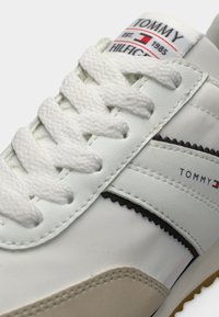 Λευκό συνθετικό αθλητικό παπούτσι με υφή, που διαθέτει λευκά κορδόνια, μαύρες ρίγες διακόσμησης και ετικέτα με λογότυπο κοντά στη γλώσσα.