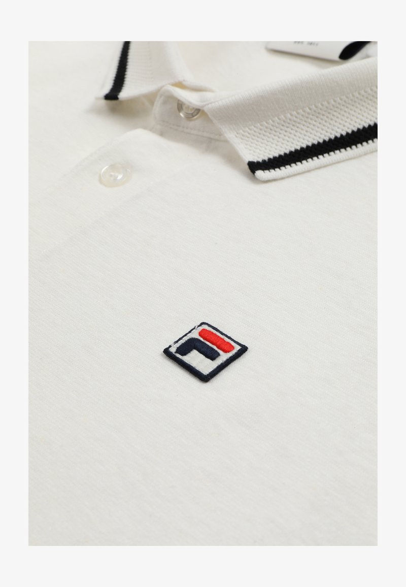 Polo blanc avec col rayé noir et blanc, boutons transparents, et petit logo carré brodé en noir, blanc et rouge sur la poitrine.