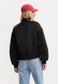 Zwarte satijnen bomberjack met opgepofte mouwen, geribbelde boorden en zoom. Gecombineerd met lichtblauwe spijkerbroek en een rode pet.