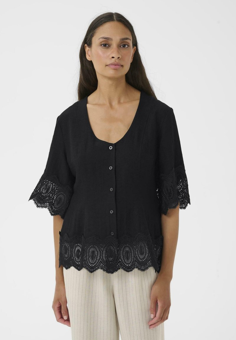 Femme portant un chemisier noir boutonné avec un bord en dentelle aux manches et à l'ourlet, associé à un pantalon beige à rayures fines, debout devant un fond uni.