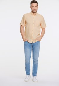 Camisa abotonada de manga corta en naranja claro con un bolsillo, combinada con vaqueros azul claro y zapatillas blancas. Diseño sencillo, ajuste informal.