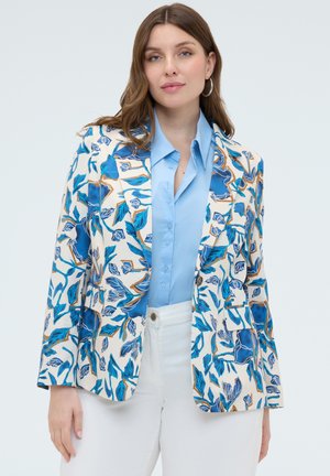 Vrouw met een blauw overhemd met knopen, witte broek en een witte blazer met blauw-bruine bloemen- en bladerenpatronen, staand tegen een effen achtergrond.