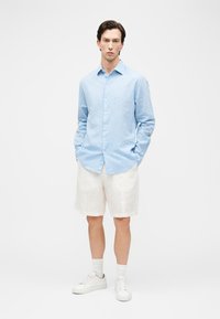 Homme debout, mains dans les poches, portant une chemise boutonnée bleu clair, un short blanc jusqu'aux genoux, des chaussettes blanches et des baskets blanches sur un fond uni.