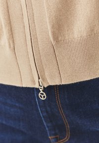 Beige kardigán cipzárral és bordás textúrával, sötét kék farmerral párosítva. A cipzárhúzó kiemelkedő, kör alakú kialakítású.