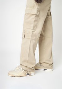 Pantalones cargo de tela beige claro con grandes bolsillos laterales, corte recto y costuras reforzadas. Combinados con zapatillas crema con detalles en negro.