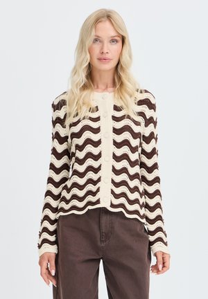 Femme blonde aux cheveux ondulés porte un cardigan en maille rayé crème et marron avec des boutons et un pantalon marron foncé, tournée vers l'avant.