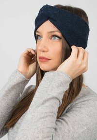 Fascia in maglia blu navy con design a torsione anteriore. Texture liscia, indossata su capelli lunghi castani, abbinata a un maglione grigio chiaro.