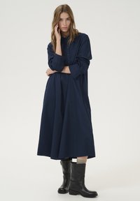 Rochie bleumarin cu nasturi, cu o siluetă lejeră și evazată. Mâneci lungi și guler. Asortată cu cizme negre până la genunchi.