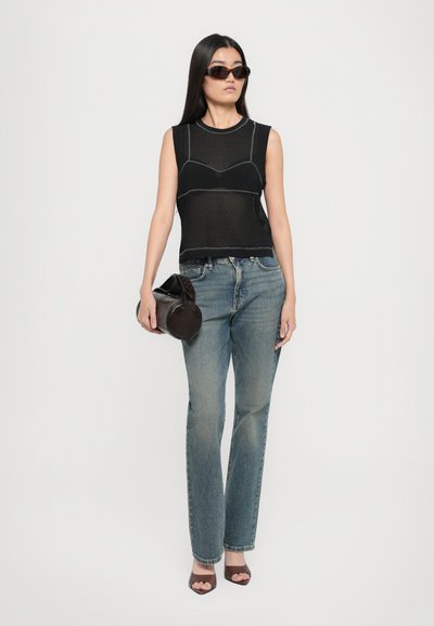 Haut sans manches noir avec des coutures contrastées, jeans bleu droit, et talons ouverts marron. Tenant un sac à main circulaire noir.