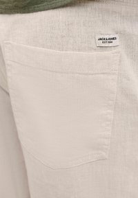 Közelkép világos bézs színű szövetnadrág hátoldali zsebével, amely felett egy fehér Jack & Jones márkajelzésű címke van varrva.