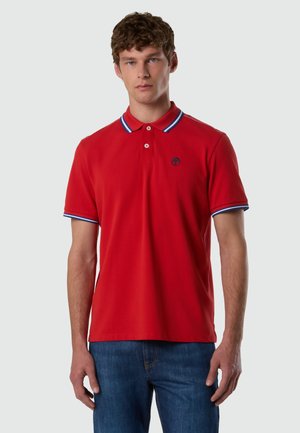 Giovane uomo che indossa una polo rossa con bordi blu e bianchi su collo e maniche, abbinata a jeans blu, in piedi davanti a uno sfondo semplice.