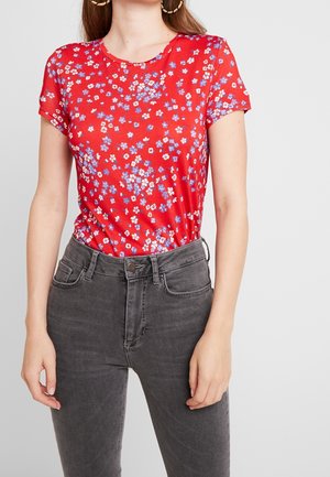 Kvinde iført rød kortærmet blomstret top stukket ind i højtaljede, falmede sorte jeans, står mod en lysegrå baggrund.