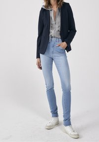 Blazer navy sobre una sudadera gris, combinado con jeans ajustados de color azul claro y zapatillas blancas. El blazer tiene un diseño estructurado y bolsillos.