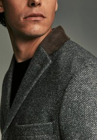Grå tweed blazer med en brun fløjlskrave, der har en notch lapel og en brystlomme. Teksturert stof med et subtilt mønster.