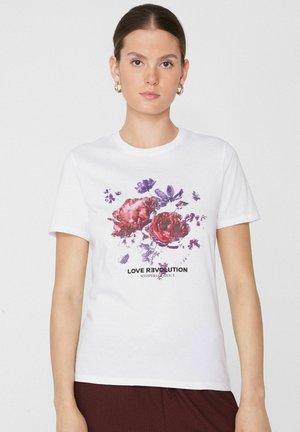 Woman with dark hair in a white T-shirt featuring red and purple flowers and text "LOVE REVOLUTION WHISPERS OF GRACE."