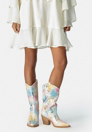 Piernas con vestido blanco con volantes que llevan botas vaqueras de textura de cocodrilo multicolor hasta la mitad de la pantorrilla con tacones gruesos sobre fondo liso.