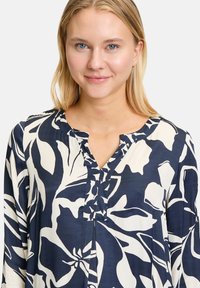 Dunkelblaue Bluse mit floralem Muster in Creme. Mit V-Ausschnitt, Knopfdetail am Kragen und langen Ärmeln. Leichte Stoffqualität.