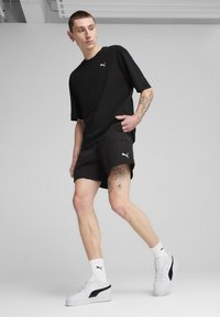 T-shirt noir surdimensionné avec un petit logo blanc, associé à un short noir avec des poches cargo, des chaussettes blanches et des baskets blanches PUMA.