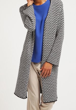 Cárdigan largo de patrón de chevron en blanco y negro, con abertura frontal, mangas largas y parte superior sólida azul a contraste debajo, combinado con pantalones beige.