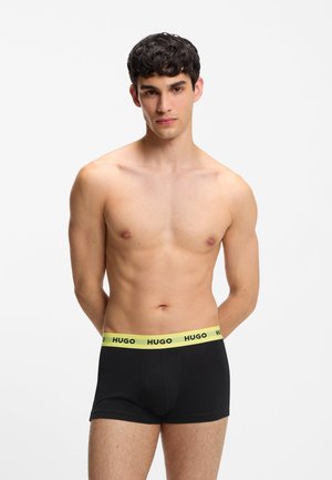 Młody mężczyzna o krótkich ciemnych włosach, ubrany w czarne bokserki z żółtym paskiem w talii oznaczone napisem „HUGO”, stojący na jednolitym białym tle.