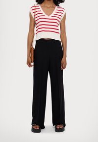 Kobieta ubrana w białą bluzkę bez rękawów w czerwone paski, czarne spodnie szerokie w nogawkach, czarne sandały na platformie oraz brązową plecioną torebkę na ramię.