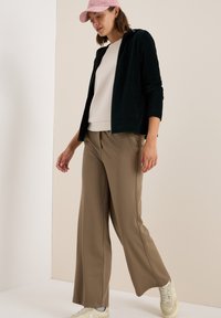 Veste zippée en velours côtelé vert foncé, sweat-shirt crème et pantalon large beige, complétés par des baskets claires et une casquette rose pâle.