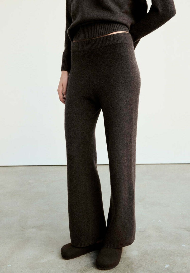 Personne portant un pantalon large en maille marron foncé et un pull assorti, debout sur un sol gris contre un mur blanc.