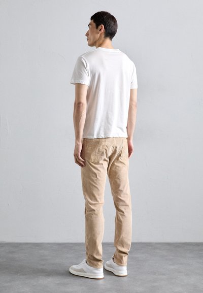 Homme debout tourné vers la gauche, montrant son dos, portant un t-shirt blanc, un pantalon beige et des baskets blanches sur un sol gris.