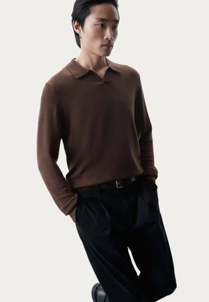 Maglione polo lungo marrone con una texture morbida, dotato di colletto a scollo, indossato con pantaloni neri, in piedi su uno sfondo chiaro.