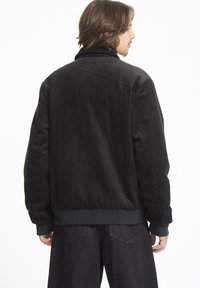 Zwarte corduroy jas met geribbelde textuur, hoge kraag en elastische manchetten. Beschikt over een relaxed fit en een ruglepdetail.