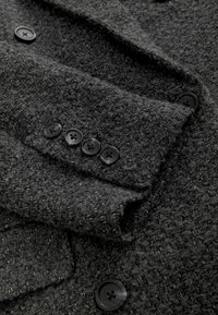 Manteau noir texturé en tissu tissé avec quatre boutons noirs aux poignets, présentant un col structuré et un léger drapé.