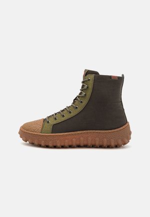 Wysokie buty z brązową teksturowaną podeszwą, oliwkowozielonym obszarem sznurowadeł oraz ciemnobrązowym materiałem wierzchnim, pokazane z boku na białym tle.