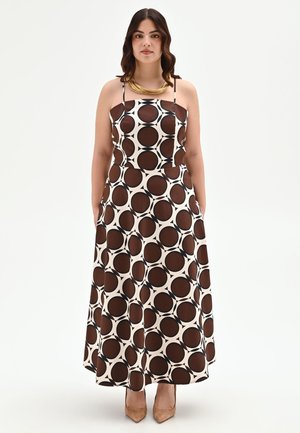 Femme aux cheveux foncés portant une robe maxi sans manches avec de grands motifs circulaires marron et blanc et des talons pointus couleur nude.