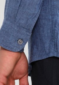 Camicia in lino blu con polsini abbottonati, cuciture verticali e orlo leggermente curvo, evidenziando una finitura di tessuto testurizzato.