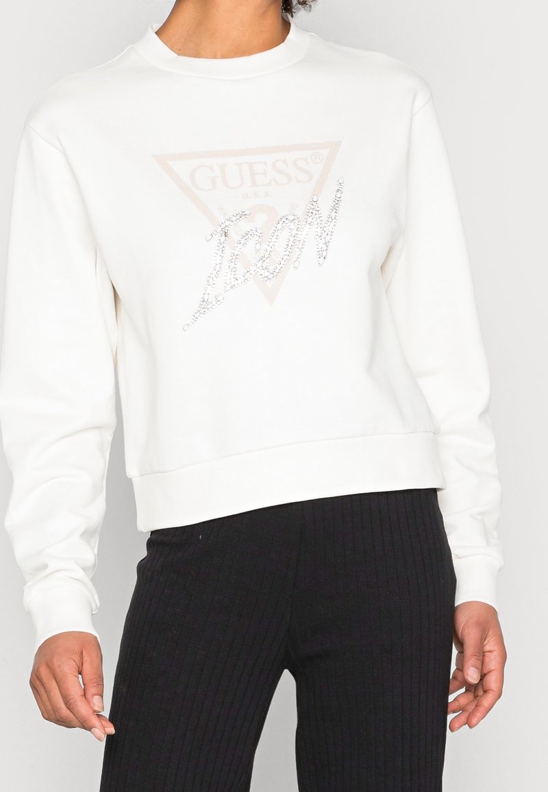 Valkoinen lyhyt collegepaita pyöreällä pääntiellä, jossa on vaaleanpunainen kolmion muotoinen logo ja kimaltava hopeinen teksti. Sileä pinta, resorireunus.