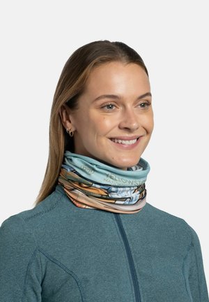 Mujer sonriendo, lleva pendientes de aro dorados, una chaqueta con cremallera de color verde azulado y un pasamontañas multicolor con el patrón del Parque Nacional Yellowstone.