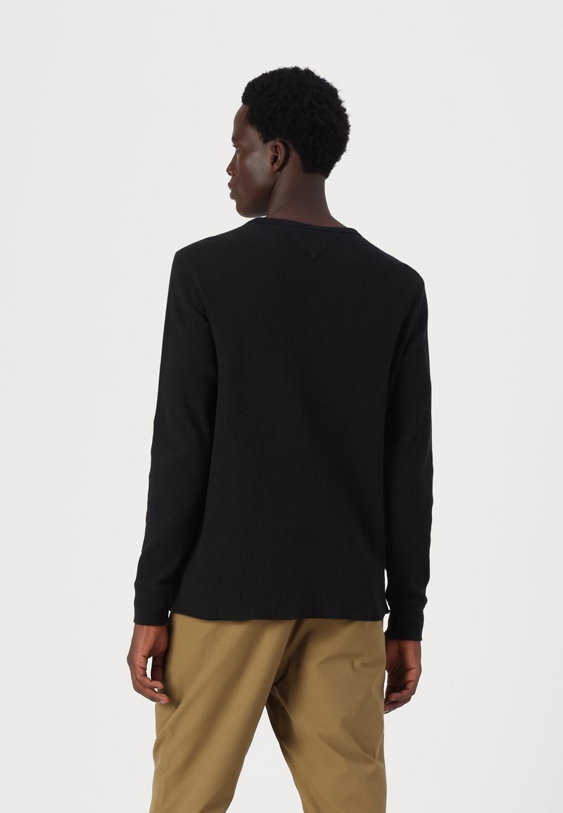 Czarny długi rękaw top o prążkowanej fakturze z okrągłym dekoltem i bocznymi rozcięciami, w połączeniu z beżowymi spodniami. Widok z tyłu pokazujący detale rękawów.