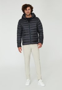 Svart pufferjacka med huva och framparti dragkedja, med kviltad yta och elastiska ärmslut, ihop med ljusbeige byxor och vita sneakers.