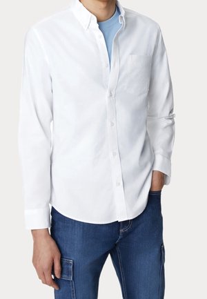 Mann trägt ein weißes Hemd über einem hellblauen T-Shirt, kombiniert mit blauer Jeans, steht mit einer Hand in der Tasche.