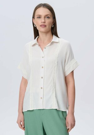 Camisa de botón ligera color beige con mangas cortas, detalles de encaje en los laterales y un corte relajado, combinada con pantalones verdes. Textura suave.