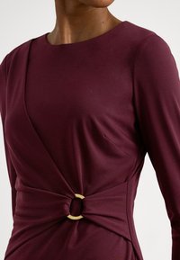 Bordo krāsu kleita ar gludu audumu, garām piedurknēm, apaļu kakla izgriezumu un viduklī ar zelta gredzenu akcentētu sasukājumu.