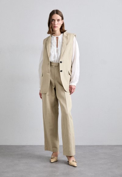 Gilet en lin beige avec de grandes poches, associé à une blouse blanche légère et un pantalon taille haute beige. Des mules à bout pointu complètent le look.
