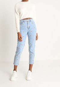 Jean en denim bleu clair avec broderie arc-en-ciel, taille haute, coupe fuselée. Associé à un pull court blanc duveteux et des baskets blanches.