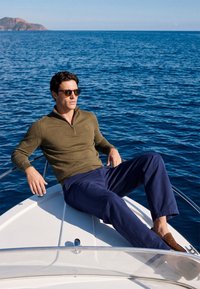 Homme portant des lunettes de soleil, un pull kaki et un pantalon bleu, allongé sur la proue d'un bateau avec un océan calme et une île lointaine en arrière-plan.
