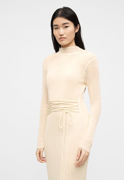 Femme aux longs cheveux noirs portant une robe en maille côtelée de couleur crème, à manches longues avec un détail de ceinture à la taille, se tenant devant un fond uni.